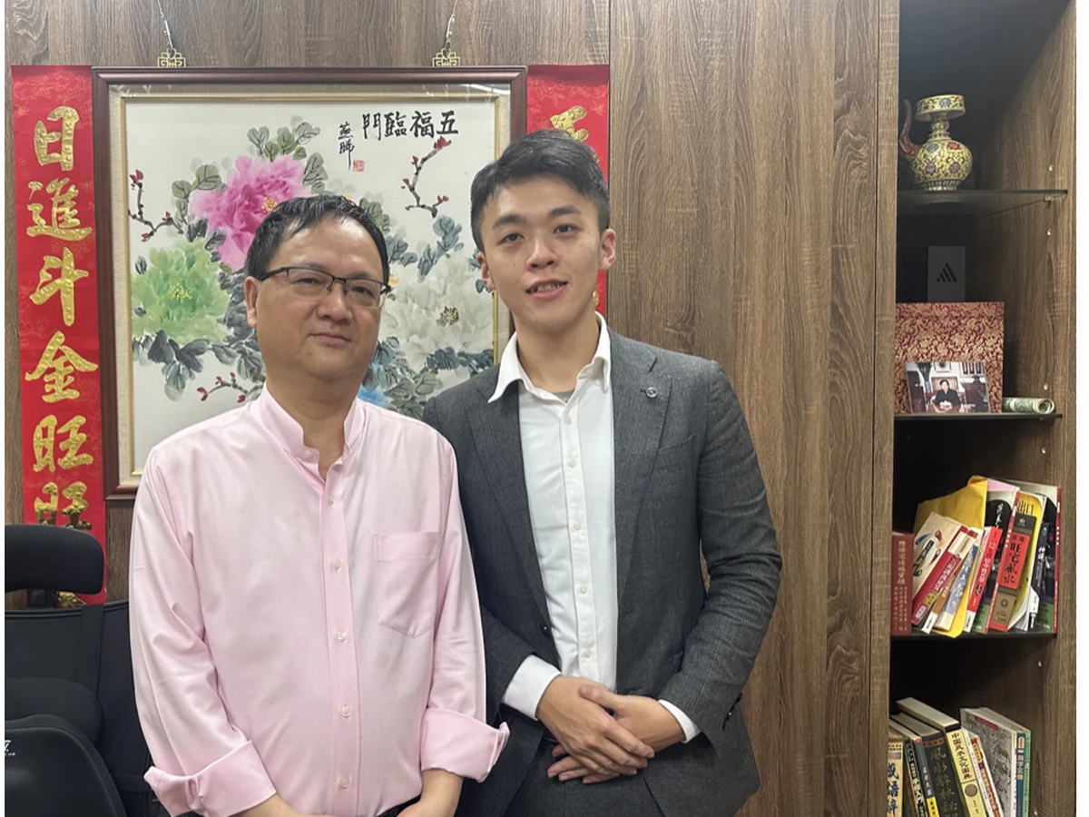 何宇軒與合作的風水命理老師合影，背景為紅色對聯、五福臨門畫作與整櫃命理書